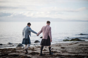 Two grooms in kilts walk hand in hand along the beach at Crear in Argyll