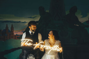 a bride and groom against a wall in oran mor holding a neon sign reading