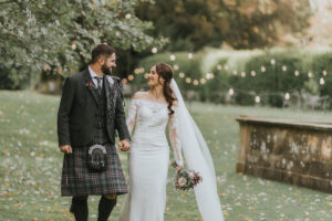 A bride wearing an off shoulder lace gown and groom wearing kilt smile at each other as they walk hand in hand over grass