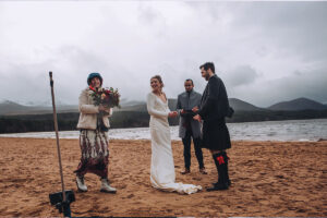 A bride and groom, celebrant and their witness stand on the shore of a loch