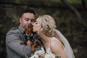 A groom holds a Dachsund dog while a bride kisses the side of the dog