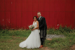 A bride and groom stand in front of a red barn wall