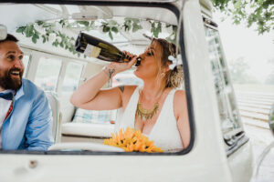 A bride and groom sit in the front of a van and the bride sips from a bottle of champagne