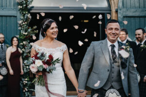 Smiling bride and groom with dark hair being showered in petal confetti with wedding guests in background