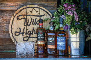 Three bottles of whisky, a silver bucket filled with flowers and two glasses filled with ice in front of a wooden sign that reads Wild Highland Drinks Co