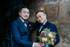 two smiling grooms on their wedding day wearing navy blue suits and carrying a bouquet of flowers