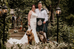 a bride and groom standing in a garden with two dogs