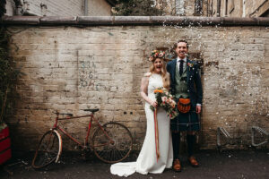 a bride wearing a flower crown and groom wearing a kilt standing next to a wall getting showered in white confetti