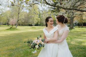 two brides hugging in the grounds of Raemoir House under a hawthorn tree with white blossom