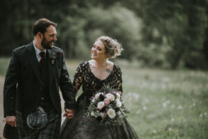 a bride wearing a black Zander wedding dress by Sottero and Midgeley holding hands with a groom wearing a kilt walking across a lawn
