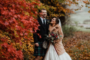 bride wearing faux fur jacket with groom in the grounds of Balbirnie House with autumn leaves