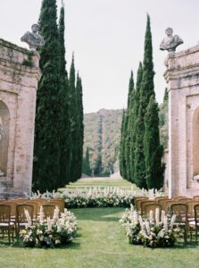 Italian villa wedding ceremony with cypress trees, cane back chairs and lush green and white flower aisle