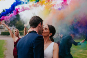 a bride and groom kissing in ground of Gilmerton House with rainbow smoke bombs in background