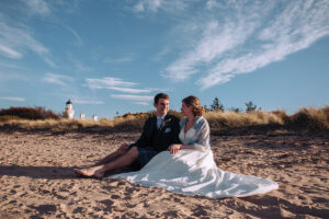 bride and groom sitting on the sand with a lighthouse in background