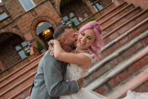 bride with pink hair and groom wearing grey kissing by the steps at Boclair House